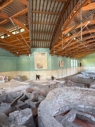 The image depicts the interior of a large, covered archaeological site in Sremska Mitrovica, Serbia. The structure features an exposed wooden beam ceiling with metal supports, and the walls are painted green and beige. The foreground is dominated by the excavated ruins of ancient brick and stone structures, with some areas showing red soil. In the background, on the walls, are large framed diagrams or mosaics.
Several human figures are visible within the ruins. On the left, there are white statues or figures in historical attire. In the center, two dark-hued statues or figures in historical dress stand together. Further in the background, another solitary figure in historical dress stands near a section of the ruins. On the right, a platform with a railing is visible, and a person in casual clothing is standing in the far distance. Red ropes are used to delineate pathways and sections of the archaeological site.