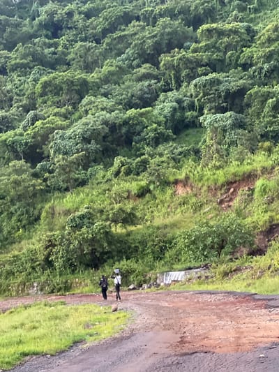 Two individuals are walking on a dirt road in a mountainous, lush green environment. The person on the left is wearing a dark outfit and carrying something on their head. The person on the right is wearing a white top and dark pants, also carrying an object on their head. The road is a mix of dirt and gravel, with some patches of grass on either side. The background is dominated by a dense, green hillside covered in trees and vegetation. A small waterfall or water feature is visible in the middle ground, flowing over rocks. The scene appears to be in Wuro Gole, Nigeria, given the context.