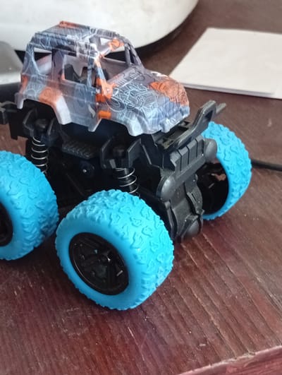 A toy monster truck with blue wheels sits on a wooden surface. The truck has a camouflage-patterned body in gray, orange, and black. Its chassis is black with visible suspension springs. The background is blurred, showing a white object and a dark surface. There is no indication of Kyshtym, Russia in the image.