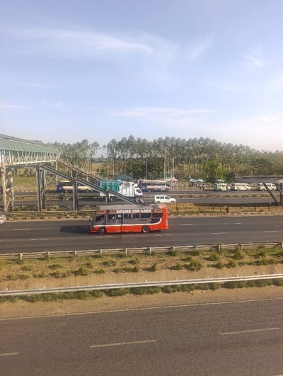 This image depicts an elevated view of a multi-lane highway in Gatongora ward, Kenya, under a clear blue sky with sparse white clouds.
In the immediate foreground, a dark asphalt surface, possibly a lower road or shoulder, is visible. It is bordered by a light grey concrete barrier with a strip of dry, patchy grass and a metallic guardrail.
The midground features a multi-lane highway, divided by a central median containing sparse vegetation and concrete barriers. Vehicles are present on the highway. A prominent red and grey "matatu" minibus, with "PRAISE THE LORD" written in white on its grey upper body, is moving from left to right in the near lane. Other vehicles, including white vans/minibuses, a light-colored sedan, and various trucks (one with a blue tarp), are visible on the highway, some moving and others appearing parked or stopped on the far right side.
To the left and spanning across the highway is a pedestrian overpass structure, constructed from metallic grey supports and featuring a covered walkway with a green corrugated roof. It includes multiple ramps leading down to ground level.
The background consists of a dense, linear stand of tall, slender trees, likely eucalyptus, stretching horizontally across the horizon. Beyond the trees, utility poles are discernible. No clearly identifiable individuals are visible within the frame.