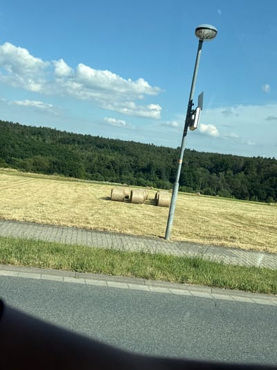 This media file shows a street-side scene in Volpertshausen, Germany. The primary objects are a metal pole with a circular light fixture at the top and a device mounted halfway down, likely a traffic camera or sensor. In the background, a field of harvested wheat, indicated by straw bales, stretches towards a dense, green forest. The scene is bordered by a paved sidewalk and an asphalt road. The sky is bright blue with scattered white clouds, suggesting a clear, sunny day, possibly late morning or early afternoon. There is no visible text in the image. The overall impression is one of a rural or semi-rural roadside setting during harvest season.