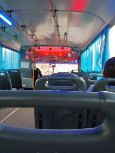 The image shows the interior of a bus in General Pacheco, Argentina. The bus is empty, with only a few seats occupied. The view is from the back of the bus looking forward. The seats are gray and have blue lights illuminating them. The ceiling of the bus has a light blue stripe running along the length. The bus is in motion, and the blur of the scenery outside the windows suggests that it is traveling at a moderate speed. There is one person, sitting in the seat on the right side of the bus, looking forward. The person is facing forward and is obscured from the view. The person is wearing a dark blue shirt and has their back to the camera. The rear of the bus has a few seats on both sides, but the front of the bus is not visible. The bus appears to be clean and in good condition. The bus has a blue curtain on the right side and a blue curtain on the left side. The bus also has blue lights on the ceiling.