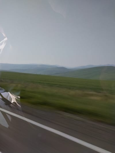 The image depicts a hazy, rolling landscape of green hills under a pale sky. The foreground shows a blurred view of a road with a white line, suggesting motion. In the reflection on the left side of the image, a person's head and shoulder can be faintly seen, wearing white clothing. The scene appears to be captured from a moving vehicle. The location is identified as Kamyshenka, Russia.