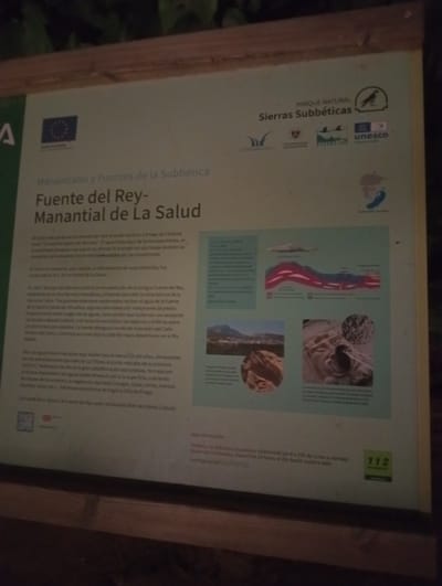 The image is a photograph of an informational sign in a park, likely a tourist attraction. The sign is in Spanish and has text and images related to "Manantiales y Fuentes de la Subbética: Fuente del Rey - Manantial de La Salud." This translates to "Springs and Fountains of the Subbética: King's Fountain - Health Spring."
The sign is located in or near Priego de Córdoba, Spain, as indicated by the text mentioning "Priego de Córdoba" and the "Villa de Priego." The environment appears to be outdoors, with a wooden frame around the sign, and some foliage is visible at the top of the image, suggesting a natural setting.
The sign's content discusses the history and geological significance of the King's Fountain and Health Spring. It mentions that the area is understood as "Ciudad del Agua y del Barro" (City of Water and Mud) and describes the origin of the water, its flow, and historical renovations. There are also several illustrative images on the sign:
* A diagram showing geological strata and water flow.
* A depiction of the landscape with hills and a town.
* A close-up of carved stone, likely from the fountain's decoration.
* A detail of the spring's water source.
Various logos are visible on the sign, including those of the "Parque Natural Sierras Subbéticas," "Unión Europea," "UNESCO," and the "Universidad de Granada." There is also a section providing contact information for "Atención Ciudadana" (Citizen Attention) and a "112" emergency number.
No people are clearly visible in the image, but the presence of an informational sign suggests a location frequented by visitors. The sign itself is the primary subject, providing information about a natural and historical site.