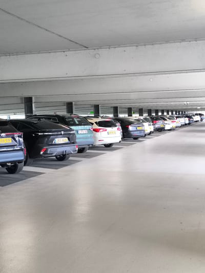 The photograph captures a brightly illuminated indoor parking garage filled with numerous cars neatly arranged in a row. The vehicles are parked between clearly marked parking spots and feature license plates with distinct characters and colors. The license plates read: "GVT-65-X", "JKF-48-B", "1-YFD-459", "26-RN". The architecture of the garage is modern with gray concrete floors, white ceilings supported by concrete beams, and linear lighting running parallel to the row of cars. The image was taken at an angle, with the camera positioned low, capturing a perspective that emphasizes the depth and organization of the parking structure. The location is Hoofddorp, Netherlands.