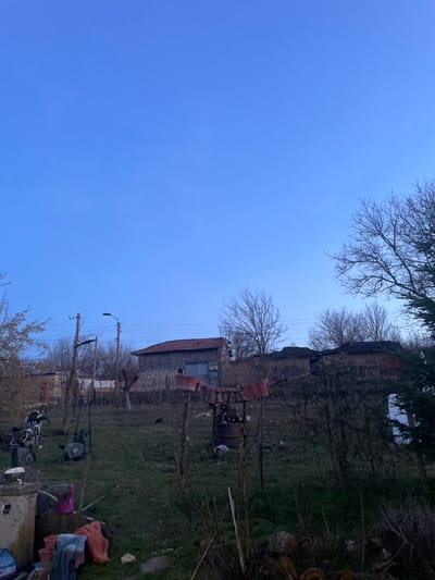 This image captures an outdoor, rural scene in Topchii, Bulgaria, under a clear, deep blue sky indicative of twilight or dawn. In the immediate foreground, a concrete wellhead with a spigot is visible, accompanied by a blue plastic bottle, a pink watering can, a red plastic container, and various fabrics. To the right, on sparse grass, a bicycle and a wheeled garden tool are positioned. Further into the mid-ground, a rudimentary structure stands, consisting of wooden posts, a metal drum at its base, and several red plastic buckets suspended from it. Utility poles with power lines and a street lamp are situated horizontally across the mid-ground. The background reveals multiple buildings on an elevated, sloping terrain: a partially obscured brick building, a two-story structure with a wooden upper section, a red-tiled roof, and a blue door, along with several older, rustic houses featuring tiled roofs. The environment is characterized by bare trees and dry bushes, suggesting a dormant season. No individuals are present in the scene.