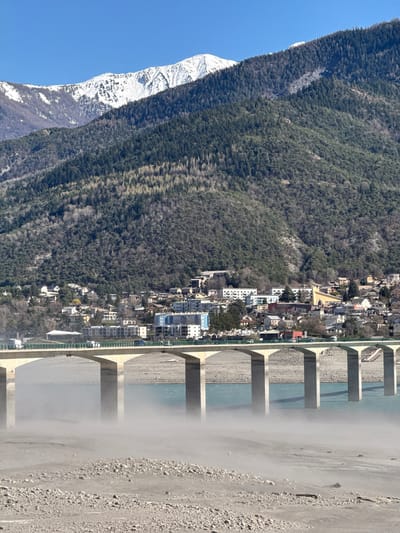 The image shows a landscape featuring a mountain partially covered in snow, a dense forest, the town of Savines-le-Lac, France, and a bridge. The town is nestled along the edge of the forest, bordering a river. The bridge spans the river and appears to be constructed of concrete. The riverbed is visible and appears largely dry. The sky is clear blue in the upper left corner. The photo seems to have been taken from an elevated vantage point.