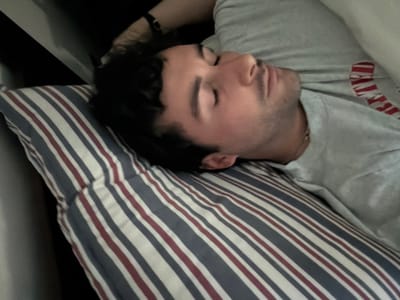 The scene, located in the city of Santo Domingo, Chile, depicts a close-up, eye-level view of a male individual sleeping. The subject, appearing to be an adult with short dark hair and light facial stubble, is lying on his right side with eyes closed, indicating a state of rest or unconsciousness. His head rests upon a rectangular pillow characterized by a pattern of alternating blue, white, and red vertical stripes. He is dressed in a light gray short-sleeved t-shirt; a red graphic or text is partially visible on the right chest area. A thin metallic chain necklace is worn around his neck. In the upper left background, a dark wristband or watch is visible on what appears to be the subject's left arm, positioned above his head. The immediate environment is dark and indistinct, suggesting an indoor setting such as a bedroom or the interior of a vehicle. No other individuals are present or interacting within the visible frame.