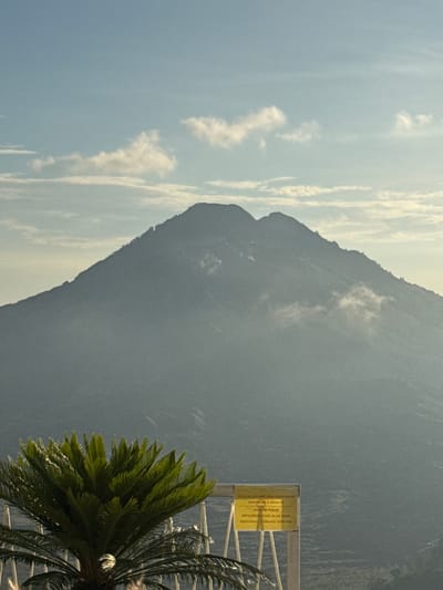 The image shows a tall, imposing mountain looming in the background. The sky is blue with scattered clouds. In the foreground, a tropical plant, possibly a palm or cycad, partially obscures a sign written in both English and Indonesian. The sign reads, "PLEASE DO NOT WALK FOOTWEAR MAKSIMUM 2 ADULTS MOHON TIDAK MENGGUNAKAN ALAS KAKI MAKSIMAL 2 ORANG DEWASA" indicating a request to remove footwear, with a maximum of two adults allowed. The location is likely Kedisan, Indonesia, based on the Indonesian language on the sign and the tropical vegetation. The scene appears to be a scenic overlook or viewing point near the mountain.
