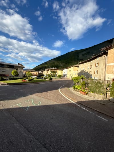 The image shows a street scene in Spormaggiore, Italy. The perspective is from ground level, looking up along a winding street lined with buildings on the left and a hill covered in greenery on the right. The sky is a vibrant blue with fluffy white clouds, suggesting a clear and sunny day. The time of day appears to be late afternoon or early evening, as indicated by the long shadows cast across the road.
The buildings are multi-story structures with a traditional European architectural style, featuring multiple windows and balconies. Some have terracotta roofs. Greenery, including trees and plants, is visible in front of the buildings and along the roadside, adding to the picturesque setting.
The road itself is paved with asphalt and has a white line marking its edge. There are chalk markings on the road, including the numbers "2" and "4", possibly indicating a temporary marking or a game. The overall impression is one of a quiet, charming Italian town bathed in warm sunlight. No people are visible in the frame. The only visible text is "MUNICIPIO" on a building in the distance, which translates to "municipality" or "town hall" in Italian.