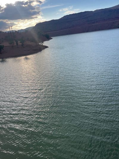 The image depicts a body of water, possibly a lake or river, adjacent to arid, rocky terrain. The water's surface is rippled, reflecting sunlight. The terrain is marked by steep cliffs and sparse vegetation. A portion of a power line infrastructure is visible along the shoreline. The location is presumed to be near Ait Mazigh, Morocco, based on the image's description, but there are no immediately identifiable features to confirm the exact location.