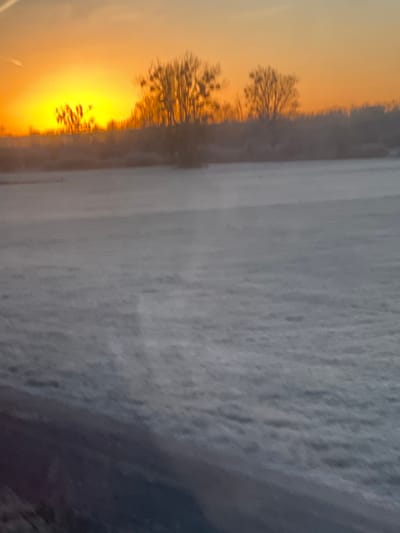 A landscape image depicts a field covered in what appears to be snow or frost. Silhouetted trees line the horizon, and a vivid orange sunrise illuminates the scene. The lighting highlights the texture of the snow-covered ground, adding depth to the composition. The image is vertically oriented.
