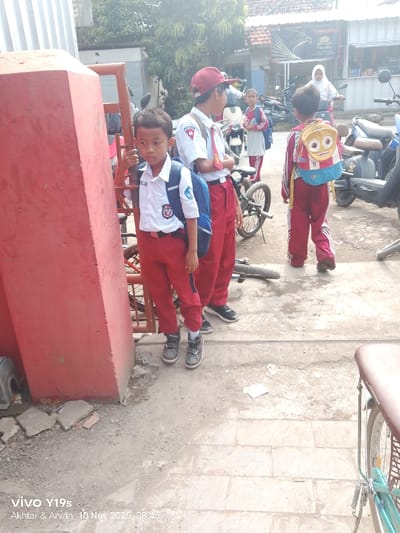 Four students in red and white uniforms stand outdoors. One boy leans against a red structure, while another wears a red cap. A third child has a backpack with a cartoon character. Bicycles and motorbikes are scattered around. A woman in a white hijab rides a motorbike in the background, near a shop. The scene appears to be outside a school or a busy street near a school.