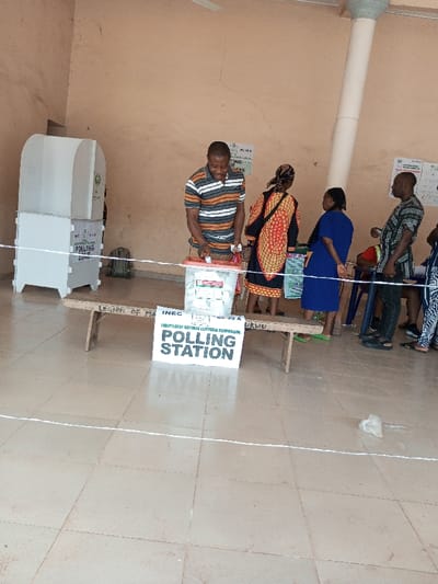 The image captures an indoor polling station in Ajalli, Nigeria, identified by an "INEC POLLING STATION" sign and a specific unit code "02-30 A UKWU". The environment consists of a light brown tiled floor, plain peach-colored walls, and a large, fluted support pillar on the right. Overhead, a ceiling fan is visible.
In the foreground, a Black male in a multi-striped polo shirt (orange, white, dark blue) is observed actively dropping a ballot paper into a transparent plastic ballot box with a red lid. This ballot box rests on a rustic wooden bench. The bench also supports a white sign displaying "INEC INDEPENDENT NATIONAL ELECTORAL COMMISSION POLLING STATION" along with the unit code. The words "LEGION OF MA" are partially visible on the wooden bench. Two white, modular polling booths are located on the far left, labeled "POLLING ZONE", with a green backpack positioned behind them.
White string lines are stretched across the tiled floor, demarcating an area. To the right of the voter, a group of four other individuals are present. These include a Black female in a patterned red, black, and yellow dress with a head covering, standing with her back to the camera; a Black female in a plain blue dress and light green sandals, standing beside her; a Black male in a dark plaid shirt, observing the scene; and partially visible individuals on the far right. Posters and notices, one displaying "ACDE C", are affixed to the wall behind the individuals. A crumpled piece of white plastic is visible on the floor in the lower right corner.