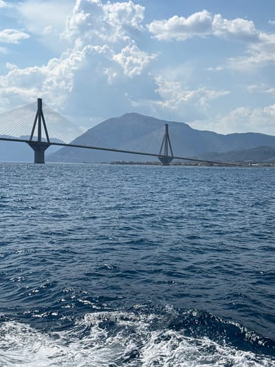 The image captures a scenic view of the Rio-Antirrio Bridge, a cable-stayed bridge spanning the Gulf of Corinth in Greece, connecting the Peloponnese peninsula to mainland Greece. The perspective is from a moving vessel on the water, with the bridge's distinctive pylons and cables dominating the left side of the frame. The deep blue water is textured with ripples and reflects the sunlight, creating a sparkling effect. White foam and spray are visible along the right edge, indicating the movement of the boat.
In the background, hazy mountains rise behind the bridge and the landmass it connects to. The sky above is a clear blue, dotted with fluffy white clouds, suggesting a pleasant day with good visibility. The overall impression is one of a journey or travel, with the impressive engineering feat of the bridge set against a beautiful natural landscape. The time of day appears to be daytime, likely late morning or early afternoon, given the brightness of the sun and the cloud formations. There is no visible text in the image.