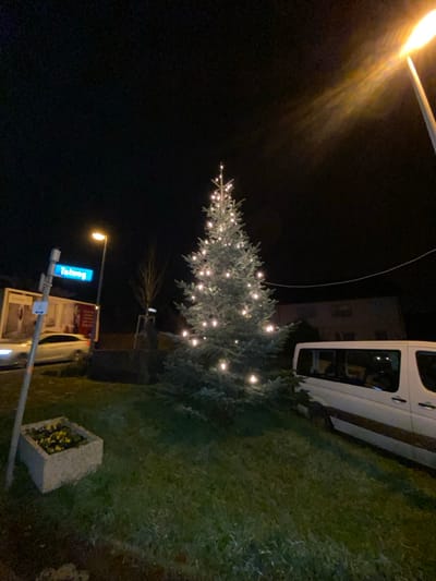 A Christmas tree adorned with lights stands near a street sign at night. A white van is parked next to the tree on a grassy area. A building is partially visible behind the van. A planter box sits on the grass as well. A lamppost casts a warm glow in the upper left of the image, contrasting with the dark night sky.