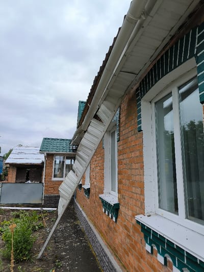 The image depicts the exterior of a brick house in Pysarivka, Ukraine, under an overcast sky. The house features light orange-brown brick walls with dark green decorative elements around white-framed windows and white window sills. A significant section of white, corrugated soffit or fascia has detached from the underside of the roof overhang, bending downwards and resting against the brick wall. A white gutter system remains largely intact along the roofline above the damaged soffit. To the left, a smaller, secondary brick structure with a green tiled roof and a lower extension, partially covered by a white tarp, is visible. The immediate foreground shows dark, damp soil with sparse green vegetation, bordering a narrow strip of gravel or small stones along the house's foundation. No individuals are present in the scene.