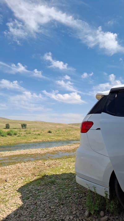 A white SUV is parked on a rocky, grassy area next to a shallow, clear river. The river flows through a dry, grassy landscape with rolling hills in the background. The sky is a bright blue with scattered white clouds. The scene appears to be a scenic overlook or rest stop in the city of Boraldai, Kazakhstan.