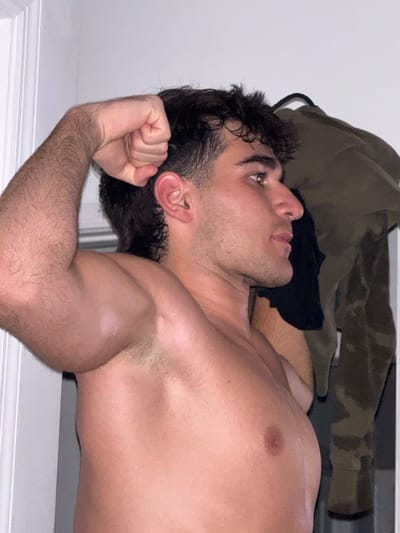 A young adult male, shirtless and with dark, curly hair, flexes his bicep while looking to the right. His left arm is raised, with his fist clenched. The background consists of a white door on the left and a rack of clothing on the right, including a green camouflage jacket. The image appears to be taken indoors, likely in a residential setting within Bellmore, United States of America.