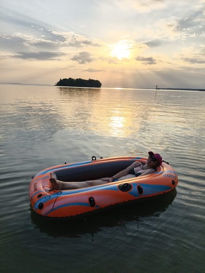 A person is relaxing in an inflatable boat on a calm body of water. The boat is orange and blue and appears to be a model named "2000". The person is wearing a bikini top, sunglasses, and a pink cap. They are lying down with their legs extended. The sun is setting, casting a warm glow on the water and clouds. In the distance, a small, tree-covered island is visible, as well as a structure that resembles a lighthouse or tower. The overall scene suggests a peaceful and serene moment on the water. The location is identified as Motala, Sweden.