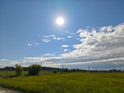 The image depicts a wide, sun-drenched grassy field under a vast blue sky with scattered white clouds. The sun is prominently positioned in the upper center of the frame, emitting a bright glare. A line of trees forms a dark green horizon in the middle ground, beyond which power lines and utility poles are visible. In the far distance, hints of buildings and structures suggest a settlement. The foreground is dominated by tall, green grass with some dried stalks, indicative of a rural or semi-rural setting. The scene appears to be outdoors during daylight hours, likely late morning or early afternoon given the sun's position and intensity.