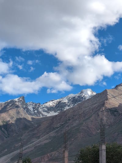The image displays a panoramic view of snow-capped mountains under a blue sky dotted with white clouds. In the foreground, unfinished concrete pillars with exposed rebar are visible, suggesting ongoing construction. The mountainous terrain is characterized by rocky slopes with patches of sparse vegetation. A notable feature is a prominent peak covered in snow, with the lower slopes exhibiting a gradient of brown and green hues. The overall scene captures a remote, high-altitude landscape.