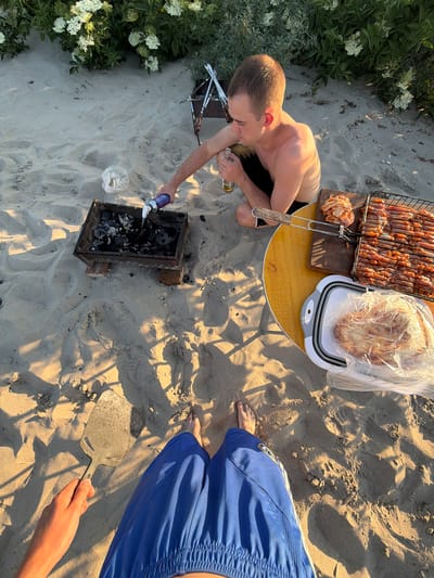 An overhead shot captures a group preparing a barbecue on a sandy beach. A man, wearing black shorts, is crouched near a portable barbecue grill, holding a bottle of beer in his right hand and a can of lighter fluid in his left, spraying it into the grill. Raw skewers of meat are on a grill rack on a yellow table to the right of the man. Next to the grill rack is a white collapsible container with raw chicken inside. In the foreground, a person's legs wearing blue shorts are visible, along with a hand holding a metal spatula. The scene is set on a beach with sand and vegetation in the background. The sun is casting long shadows, indicating it is either early morning or late afternoon. The location is identified as the city of Поваровка, Russia.