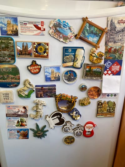 This media file is a close-up shot of a refrigerator door adorned with numerous magnets. The magnets depict a variety of subjects, including landmarks, food and drink items, cartoon characters, and abstract designs. Several magnets appear to be souvenirs from different countries, indicated by text in various languages such as Russian, Turkish, and what looks like Spanish or Italian.
Specific objects include magnets shaped like buildings (e.g., Gaudi Barcelona, St. Petersburg, Alanya), food items (e.g., Paulaner beer, Havana Club), a wooden-framed picture, a bicycle, a Santa Claus figure, and a collection of advertising magnets. Some magnets have text describing services like "Ремонт" (Repair) in Russian, offering repairs for household appliances and plumbing. Others advertise places or brands like "KRUŠOVICE" and "AMSTERDAM."
There are no visible people in the image. The setting is clearly an interior, likely a kitchen, given the refrigerator. The lighting suggests it is daytime. The overall impression is one of collected memories and personal mementos. The magnets are densely packed on the refrigerator door, creating a colorful and busy collage.