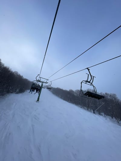 The image captures a winter scene from the perspective of someone riding a ski lift at a ski resort in Jay, United States. Several people are visible sitting in the ski lift chair immediately below the camera's position, heading up the snowy mountain.
The primary subjects are the ski lift chairs and cables, which dominate the upper portion of the frame, and the snow-covered ski slope stretching out below and to the right. The lift line ascends a wide, treeless piste, flanked on both sides by dense, bare deciduous trees, indicative of a winter landscape. The sky is a pale, overcast blue-grey, suggesting either early morning, late afternoon, or a heavily clouded day. Light snow flurries appear to be falling, visible as small white specks against the sky, contributing to the wintry atmosphere.
The activity taking place is recreational skiing or snowboarding, with individuals being transported uphill by the chairlift. The snow on the slope shows signs of recent grooming or skier traffic, with some smooth sections and gentle undulations. The image is tilted, giving a dynamic, diagonal composition to the ski lift cables and the horizon line where the slope meets the trees. There is no visible text in the image.