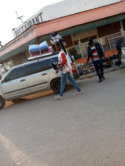 The image depicts a street scene in Ildamat ward, Kajiado, Kenya, outside the Moigucu Hotel Bar & Restaurant. Natural daylight illuminates the scene, casting shadows towards the lower left.

In the midground, a light-grey station wagon is parked facing left, equipped with a roof rack on which two blue and purple rolled items are secured. A male individual walks in front of the vehicle from left to right, wearing a white and red varsity-style jacket, blue jeans, and grey slip-on shoes with three dark stripes. He carries a bundle of multi-colored garments in his left arm and balances a large, tall stack of folded clothing on his head.

Behind this individual and to the right, another male subject walks in the same direction. He is attired in a black baseball cap, a dark blue jacket over a horizontally striped shirt, dark trousers, and brown lace-up shoes. A third individual's lower body is partially visible on the far right.

The background features a building with a peach-colored lower facade and a white upper section. Prominent signage reads "RESTAURANT" and "MOIGUCU HOTEL BAR & RESTAURANT." A green striped awning is visible below the signage. A decorative metal fence lines the front of the building, behind which numerous pairs of shoes are displayed on the ground. A satellite dish is mounted on a pole above the building. The street surface is a light-colored, unpaved or lightly paved material.
