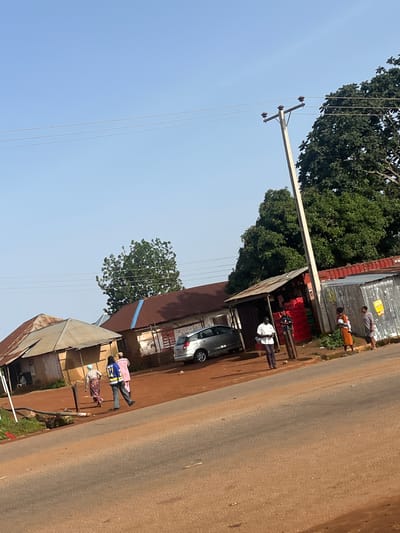 In Kassa, Nigeria, a dirt road runs alongside a few small shops and residential structures. The shops appear to be informal kiosks with corrugated metal roofs and open fronts. One shop displays red crates filled with goods. Several people are visible on or near the road. Two individuals, one in a pink garment and the other in a patterned skirt and headscarf, walk away from the camera on the dirt path. Another person, wearing a blue and yellow reflective vest over dark pants, walks alongside them. Further down the road, three more people stand outside a shop. One person, wearing an orange dress, holds a baby. A silver car is parked in front of one of the structures. A utility pole with wires stretches across the scene, with a large tree providing shade in the background. The sky is clear and blue, suggesting a sunny day. The overall impression is of a busy, albeit modest, commercial and residential area in a Nigerian town.