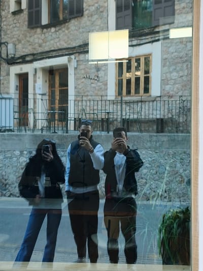 This image depicts a reflection in a window, revealing three individuals engaged in taking photographs of their reflection or selfies, superimposed over a view of a street and stone buildings in Fornalutx, Spain.
From left to right, the reflected individuals are:
1. A person with long, dark, wavy hair, wearing sunglasses on their head, a dark jacket over a light-colored top, and blue jeans. They are holding a dark smartphone in their right hand, pointed outwards.
2. A male, wearing sunglasses on his head, a dark-colored padded vest over a long-sleeved white top, and dark pants. He has a ring on his right hand and a bracelet. He is holding a dark smartphone in both hands, pointed outwards.
3. A male, wearing a dark jacket over a white top and dark pants. His face is largely obscured by the dark smartphone he is holding in both hands, pointed outwards.
The background, visible through the window, features traditional stone buildings characteristic of Fornalutx. These structures exhibit irregular, light-colored stone facades. One building displays multiple windows with dark-brown wooden shutters, some closed, others open. A doorway with a wooden door is also visible. Below the buildings, a stone wall supports a dark metal railing. Behind this railing, several dark metal tables and chairs are arranged, indicative of an outdoor seating area. Overhead wires are visible against the stone facade.
Other elements include a prominent rectangular light fixture reflected in the upper center of the window and a green potted plant partially visible in the lower right corner, suggesting it is located inside the establishment from which the photograph was taken. The street or pavement in front of the buildings is dark.