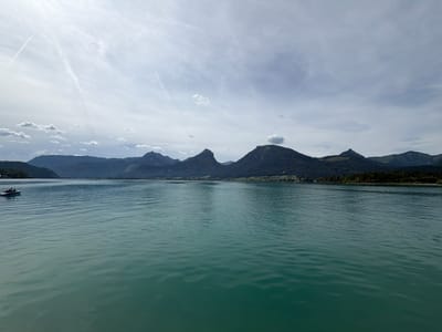 A wide shot captures a serene lake with a distant mountain range under a partly cloudy sky in St. Wolfgang im Salzkammergut, Austria. The turquoise water is slightly rippled, reflecting the sky and mountains. In the foreground, the water fills the lower half of the frame. A small white boat with two people on board is visible on the left side of the image, moving away from the viewer. The middle ground shows the tree-covered slopes of the mountains, with some scattered houses and fields visible. The background features the jagged peaks of the mountain range, some with cloud formations around them. The sky is a mix of white clouds and blue, with faint contrails from an aircraft. The overall impression is one of natural beauty and tranquility.