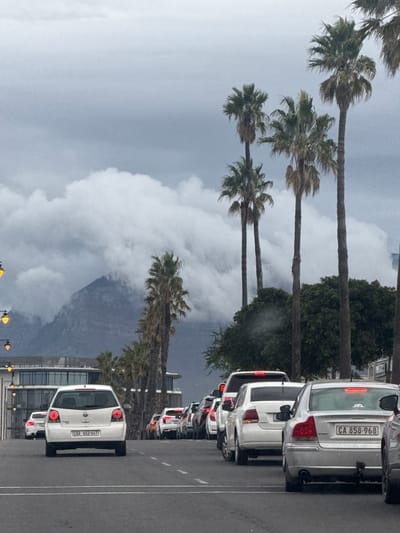 The image shows a street in Milnerton, South Africa. On the right is a line of parked cars on the side of the road, and palm trees line the left side of the street. In the background, there are clouds over a mountain. The sky is overcast.