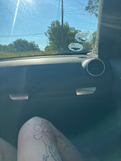 The image captures a sunlit interior view from a car, looking out the passenger side window and across to the vehicle's interior. On the far right, the light-skinned arm and shoulder of a person are visible, featuring a prominent tattoo of a flower, outlined in a reddish-purple hue, along with other grey/black tattoos.
The car's interior, visible on the left and upper parts of the frame, includes a circular air vent in the upper-left quadrant and a metallic, vertically oriented grab handle or trim piece in the mid-right. The side window, appearing somewhat streaky with dust or water marks, showcases bright sun glare. Affixed to the window are two partially peeled, circular stickers; one is mostly residue, while the other clearly depicts a white circle containing a black cross or plus sign symbol.
Through the window, a clear blue sky is visible, along with green trees and foliage suggesting a natural or rural roadside environment. Thin power lines and a utility pole are also discernible against the sky. The bright illumination indicates it is daytime with clear weather. The scene is set in Rogozen, Bulgaria. No specific activity is occurring, and there is no legible text on any visible objects, apart from the cross symbol on the sticker.