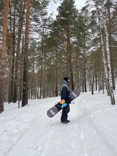 In a snow-covered forest in Sliznevo, Russia, a male individual stands facing right, holding a snowboard. He is dressed in a two-tone black and gray winter jacket, black pants, dark boots, and a black balaclava covering his head and lower face. The snowboard, held vertically with its nose upwards, is gray with "ChilDom" branding. It features black bindings with light blue accents, and a colorful graphic design in shades of pink, yellow, blue, and black is visible on its underside near the bindings. The individual stands on a snowy path marked with visible tracks, surrounded by numerous tall pine trees and several birch trees, under an overcast sky.