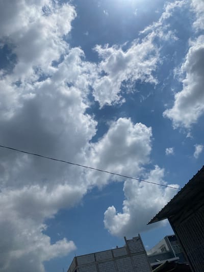 A low-angle shot captures a bright, sunny sky filled with fluffy white cumulus clouds against a vibrant blue backdrop. The sun is visible as a bright white orb in the upper right quadrant of the image, casting a strong light. A thin black wire stretches horizontally across the middle of the frame. In the lower portion of the image, the dark, corrugated metal roof of a building is visible on the right, angled upwards. To the left of the roof, the top section of a white brick building under construction is visible, with exposed rebar at the top. Further to the left and behind the brick building, a modern white and gray multi-story building with windows can be seen. The scene suggests a daytime view from a partially urban or developing area.
