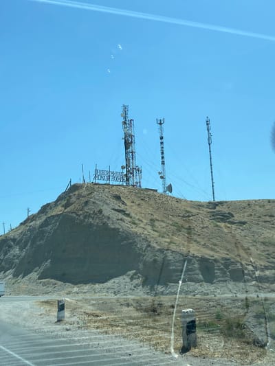 The image shows a dry, arid landscape with a hill in the background. On top of the hill are several telecommunication towers and a large sign with text. The sign appears to be in Cyrillic script. The environment is sunny with a clear blue sky. In the foreground, there is a road with white lines and a small concrete marker on the side. The overall scene is likely a rural or semi-urban area. Given the context of Kurkat, Tajikistan, this could be a viewpoint or a location with significant infrastructure.