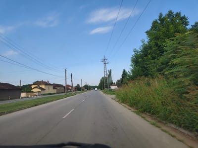 The image shows a view from a vehicle traveling along a road in Letnitsa, Bulgaria. The road is paved and has a white line marking the lane. On the left side of the road, there are several houses with tiled roofs and a grassy verge. On the right side, there is a tall tree and a dense field of tall grass. Overhead, power lines crisscross the sky, supported by utility poles and a large lattice tower. In the distance, a white van is visible on the road. The sky is a clear blue with a few wispy clouds.