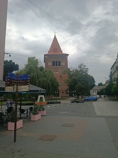 This is a medium-shot, eye-level, horizontal, outdoor photograph taken on an overcast day in Drohobych, Ukraine. The scene depicts a street with a prominent brick tower building on the left, a street with buildings on the right, and a paved public area in the foreground.
The brick tower building, likely a church or historical landmark, features a red-tiled pyramidal roof topped with a cross. It has arched windows on its upper level. To the left of the tower, a pink building is partially visible. In front of the tower, a large weeping willow tree is prominent.
A signpost with multiple directional signs in Ukrainian is situated to the left of the tower. Some signs indicate distances and names of streets and landmarks, such as "вул. Задвірня," "вул. Трускавецька," "Церква св. Юра," and "Мiська полiклiнiка." Below the signpost is an open-air seating area with tables and chairs, covered by a dark-colored umbrella or awning. Two women are walking past the seating area.
The foreground is paved with cobblestones. Pink rectangular planters containing green plants are placed in front of the seating area. In the middle ground, a blue SUV is parked on the street. The street on the right is also paved with cobblestones and lined with buildings. The sky is heavily clouded.