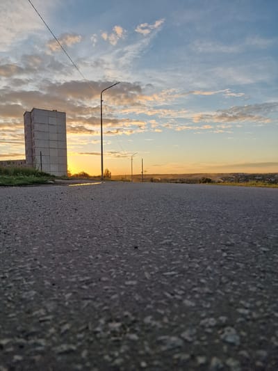 A low-angle shot shows an asphalt road in the foreground, with a tall, grey, multi-story building to the left. A utility pole with a light fixture stands near the building, and another pole is visible further down the road. The sky above is a mix of blue and scattered clouds, with the sun setting on the horizon, casting an orange glow. In the distance, a town or city is visible, with houses and trees. The scene appears to be outdoors in an urban or suburban environment.