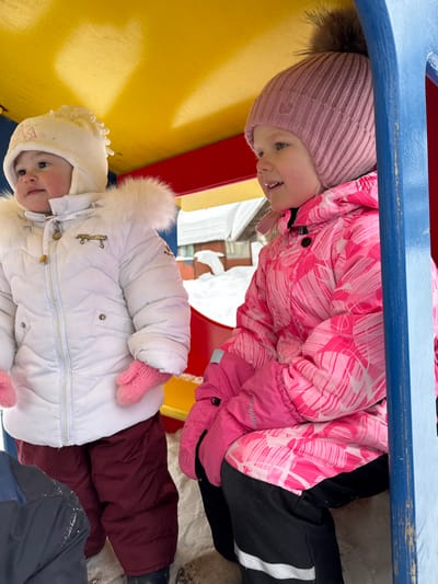Two young children are observed within a brightly colored outdoor playground structure in Zaosinovo, Russia, during what appears to be a snowy winter day. The structure features a yellow ceiling, red side panels, and blue vertical supports.
The child on the left, a younger individual, stands facing slightly left, looking upward with a serene expression. This child wears a white quilted winter coat with a fur-trimmed hood, a white knitted hat with ear flaps, and pink knitted mittens. Dark red snow pants are visible below the coat, and dark footwear is partially obscured by snow on the ground.
The child on the right, an older individual, is seated and positioned towards the right, looking out of frame with a slight smile and open mouth. This child wears a pink winter jacket with a white abstract pattern, a ribbed pink knitted hat topped with a large, fluffy brown faux fur pom-pom, and bright pink winter gloves. Black snow pants with white reflective stripes near the cuffs complete the attire.
The ground within the structure and in the background is covered with snow. Beyond the structure, a building with a white, snow-covered roof is partially visible, indicating a potentially residential or rural setting. The lighting is bright and diffused, consistent with an overcast winter environment.
