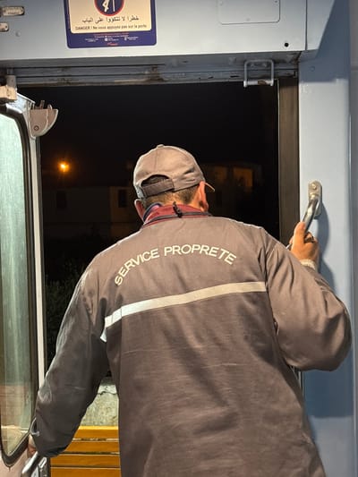 A man in a gray uniform with "SERVICE PROPRETE" printed vertically on the back opens a train door. He wears a gray cap and a face mask. The train carriage is blue, and a warning sign in Arabic and French reads "DANGER! Ne vous appuyez pas sur la porte." The exterior is dark, with a distant street light visible through the open door. A handle is attached to the door.
