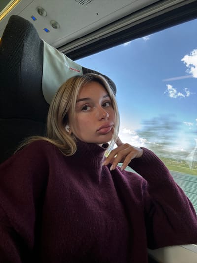 The image shows a young woman seated in a train car. She is wearing a dark purple sweater and has her left hand resting on her chin. She appears to be looking off to the left side of the frame. The window beside her shows a blurred view of the passing landscape and blue sky with clouds. There is a black headrest behind her, partially obscured by a grey headrest cover that reads "ÖBB" in red.  There are two buttons with blue lights above the headrest. The coordinates provided correspond to the town of Enns in Austria, but the specific location within the town cannot be determined from the image.  The train car appears to be modern and clean. The overall tone of the image is casual and intimate.