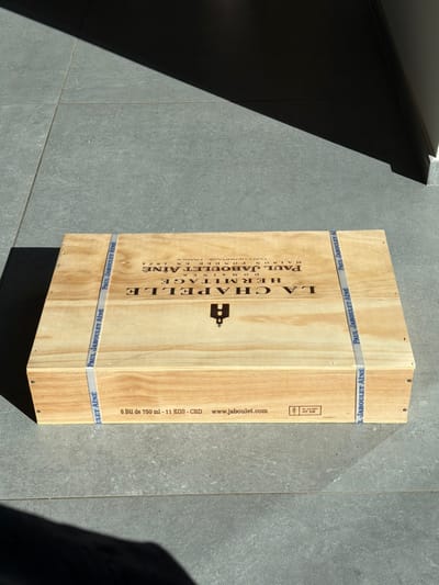 A light-colored wooden wine crate is depicted resting on a gray tiled floor. The precise location within Marche-lez-Écaussinnes, Belgium, cannot be ascertained from the image's content. The crate's top lid features black text including "LA CHAPELLE," "DOMAINE HERMITAGE," "PAUL JABOULET AÎNÉ," "MAISON FONDÉE EN 1834," and "TAIN L'HERMITAGE - FRANCE," alongside a stylized chapel icon. Two pale blue-and-white straps, marked "PAUL JABOULET AÎNÉ," are secured around the crate, running across its width. The visible long side panel of the crate displays "6 Btl de 750 ml - 11 KGS - CRD," "www.jaboulet.com," and an "HT DB" stamp. The floor is composed of large, square, unpolished light gray tiles with dark grout lines. Intense, directional sunlight illuminates the scene from the left, casting a prominent sharp, dark diagonal shadow across the upper right quadrant of the image and a partial shadow in the lower left. No individuals or other dynamic subjects are present.