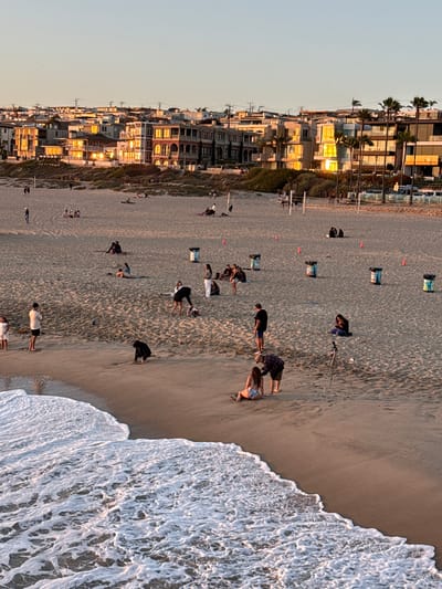 This is a medium-angle, full shot of a beach in Manhattan Beach, California, during sunset. The warm light of the setting sun illuminates the buildings in the background, casting long shadows across the sand. The beach is populated with people engaged in various activities. Some are walking, some are sitting and relaxing, and others appear to be posing for photographs. The ocean is visible in the foreground, with gentle waves lapping at the shore. The scene evokes a sense of calm and leisure, typical of a beachside community at the end of the day.