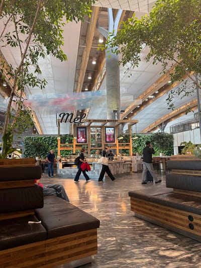 This is a photograph of the interior of a modern airport terminal in Gardermoen, Norway. The space features a high, arched ceiling with exposed wooden beams and a translucent roof allowing natural light to filter in. Various trees with green foliage are strategically placed throughout the area, some reaching towards the ceiling.

In the mid-ground, there is a food and beverage establishment with a sign that reads "Mel Norwegian Bakery." The establishment has a vertical green wall as its backdrop, and wooden shelving displays various products. A counter with display cases filled with food items is visible.

Several people are present in the scene. To the left, a man in a blue shirt and jeans is standing near the counter, appearing to be an employee. Further to the left, a person is seated on a bench, partially visible. In the center of the image, a woman in a black top and dark pants walks by, carrying a red jacket and a bag. To the right of her, a man in a dark jacket and grey pants walks with a suitcase. Another person is seen in the far right, walking away from the camera.

The floor is a polished, marble-like surface with a marbled pattern. Seating areas with dark leather benches and wooden bases are visible in the foreground and to the right. The overall atmosphere suggests a busy, yet well-designed, public space.
