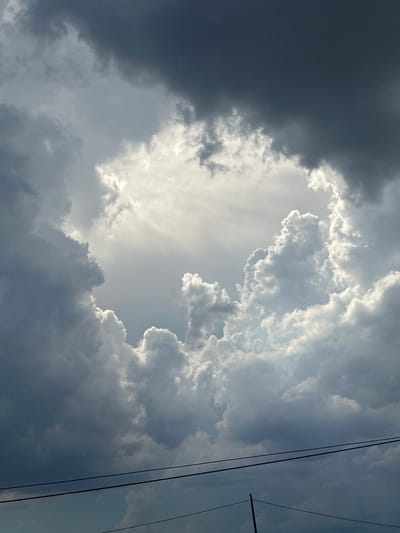 The image depicts a dramatic sky filled with dark, heavy storm clouds and bright, illuminated clouds, creating a contrast that forms a rough, oval-shaped opening. The bright clouds are puffy and textured, catching the sunlight which streams through the opening, illuminating the scene from above. The dark clouds surround this bright area, appearing dense and foreboding. In the foreground, two thin, dark wires run horizontally across the lower portion of the image, suggesting the presence of a power line or telephone line. Below the wires, a thin, vertical pole is visible on the right side of the frame. The overall impression is of an impending storm with a break in the clouds allowing sunlight to pierce through. The image does not contain any elements that would specifically place it in the city of Assisi, Italy; it is a generic sky scene.