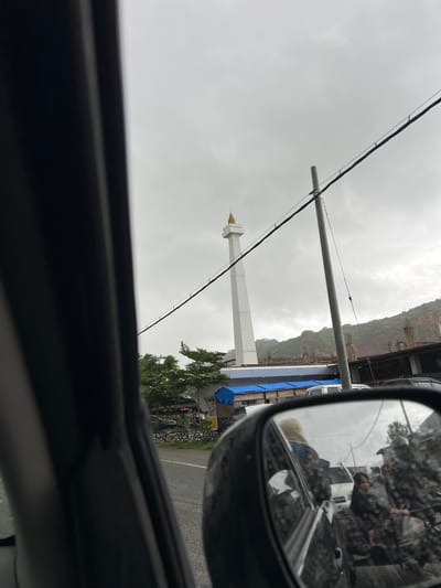 The image, taken from inside a vehicle, depicts a cloudy day in the city of Keuneueu, Indonesia. In the distance, a tall, white tower with a golden spire stands against the overcast sky. A power line runs diagonally across the frame, supported by a grey utility pole. In the foreground, the interior of the car is visible, including the driver's side window and the side-view mirror. The mirror reflects a street scene with several people on motorcycles, some wearing camouflage-patterned clothing and helmets. Buildings and trees line the street, with a hillside visible in the background. Construction appears to be ongoing at a building near the base of the tower, with scaffolding and blue tarps present.