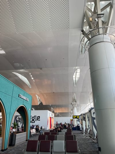 The image depicts the interior of an airport in Ramunia Dua, Indonesia. A Starbucks store with a green facade and prominent signage is in the foreground. Rows of chairs are situated near the Starbucks, with people seated and waiting. A store labeled "ets giggle" is also present. A sign indicates "Pintu/Gate 8." The architectural elements include high ceilings, a large cylindrical structure, and walls with a perforated pattern. Natural light is present, entering through windows and highlighting the interior.
